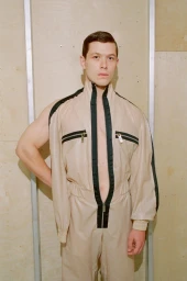 Model wearing a zipped beige jumpsuit with black trim against a light wood panel background.
