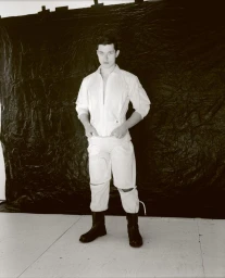 Man wearing a white jumpsuit and black boots standing against a crinkled dark backdrop in a studio setting
