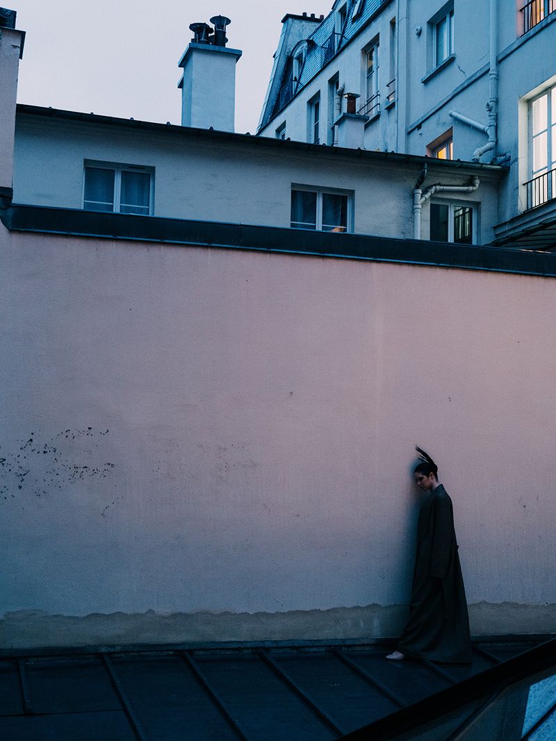 A person in a long robe stands against a tall pink wall in an urban setting at dusk, with buildings in the background.