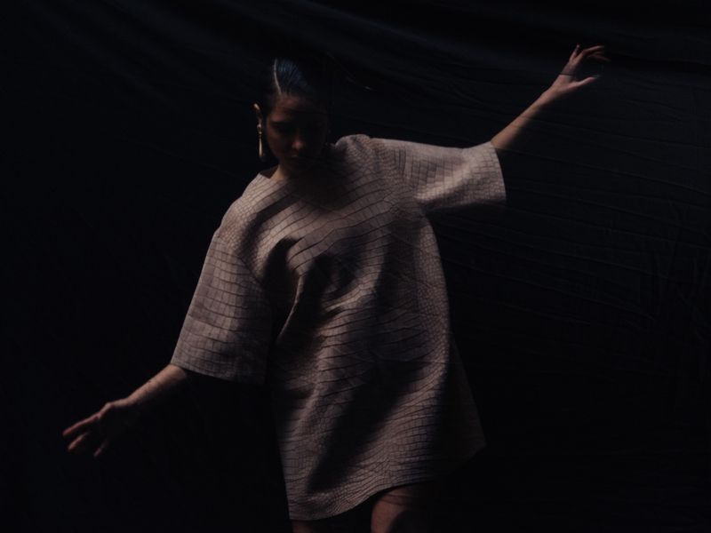Woman in a structured light-brown dress stands with arms outstretched in a dimly lit space with a draped backdrop and dark atmosphere.