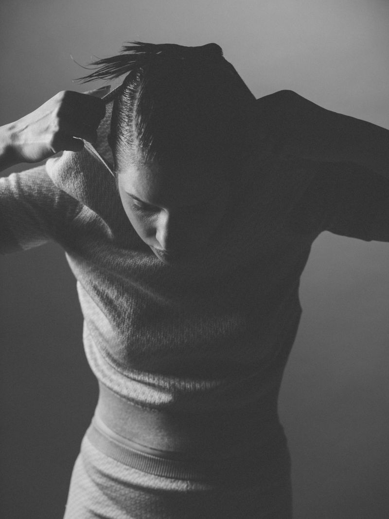Monochrome portrait of a woman adjusting her hair with both hands, downward gaze, neutral expression, against a blurred grey backdrop.