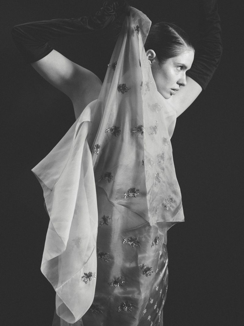 Elegant portrait of a woman in long gloves holding a sheer, embellished shawl against a dark backdrop in a monochrome aesthetic.