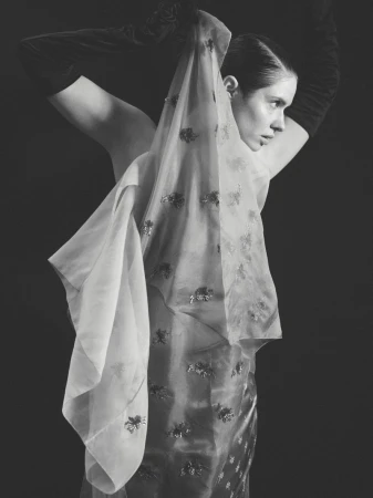 Elegant portrait of a woman in long gloves holding a sheer, embellished shawl against a dark backdrop in a monochrome aesthetic.