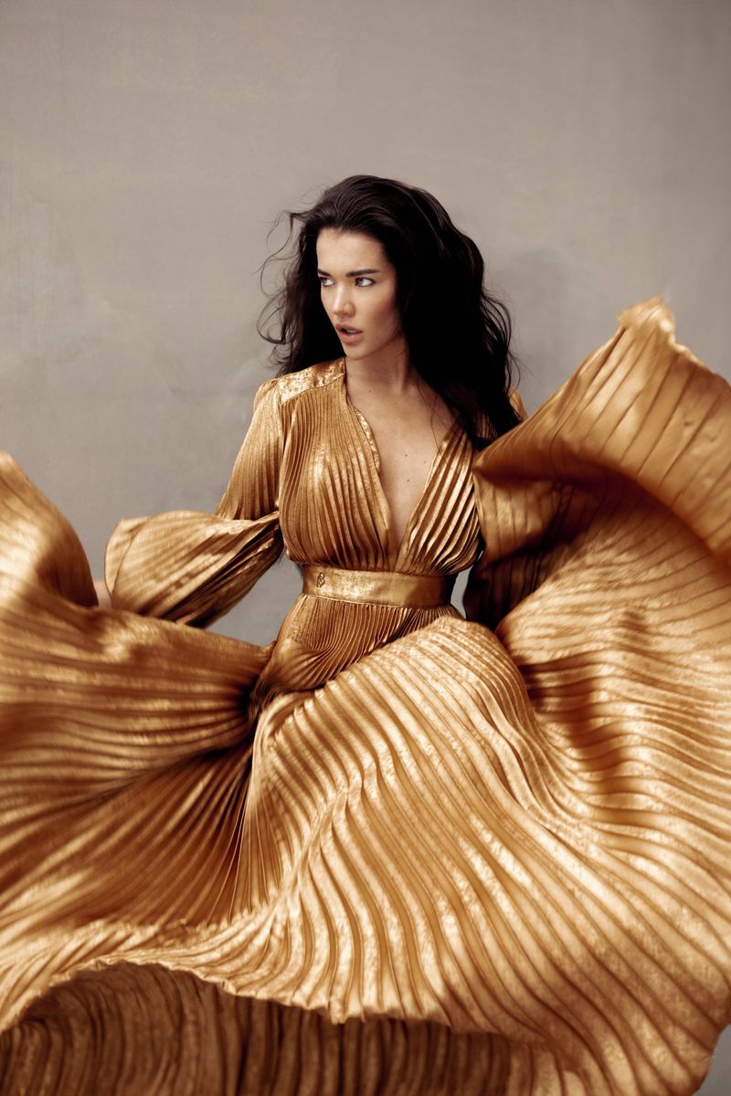 Elegant brunette woman in a flowing, golden pleated dress against a neutral background. Fashion portrait with dynamic movement.