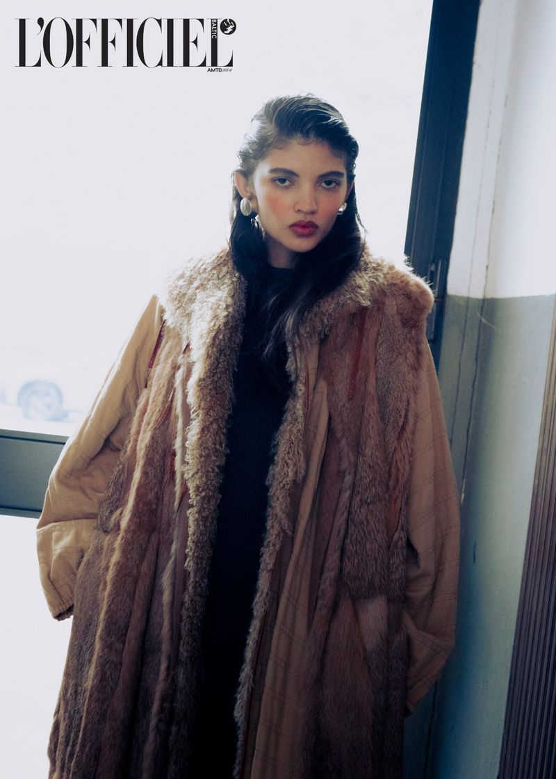 Portrait of a young woman wearing a fur-lined coat and large earrings standing indoors near a window with natural lighting.
