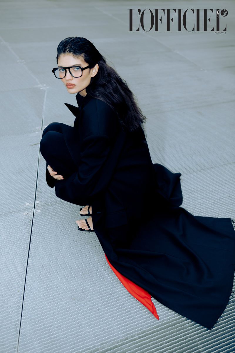 Dark-haired model in a black coat and square glasses squats on a grey platform for a fashion editorial in L'Officiel magazine.