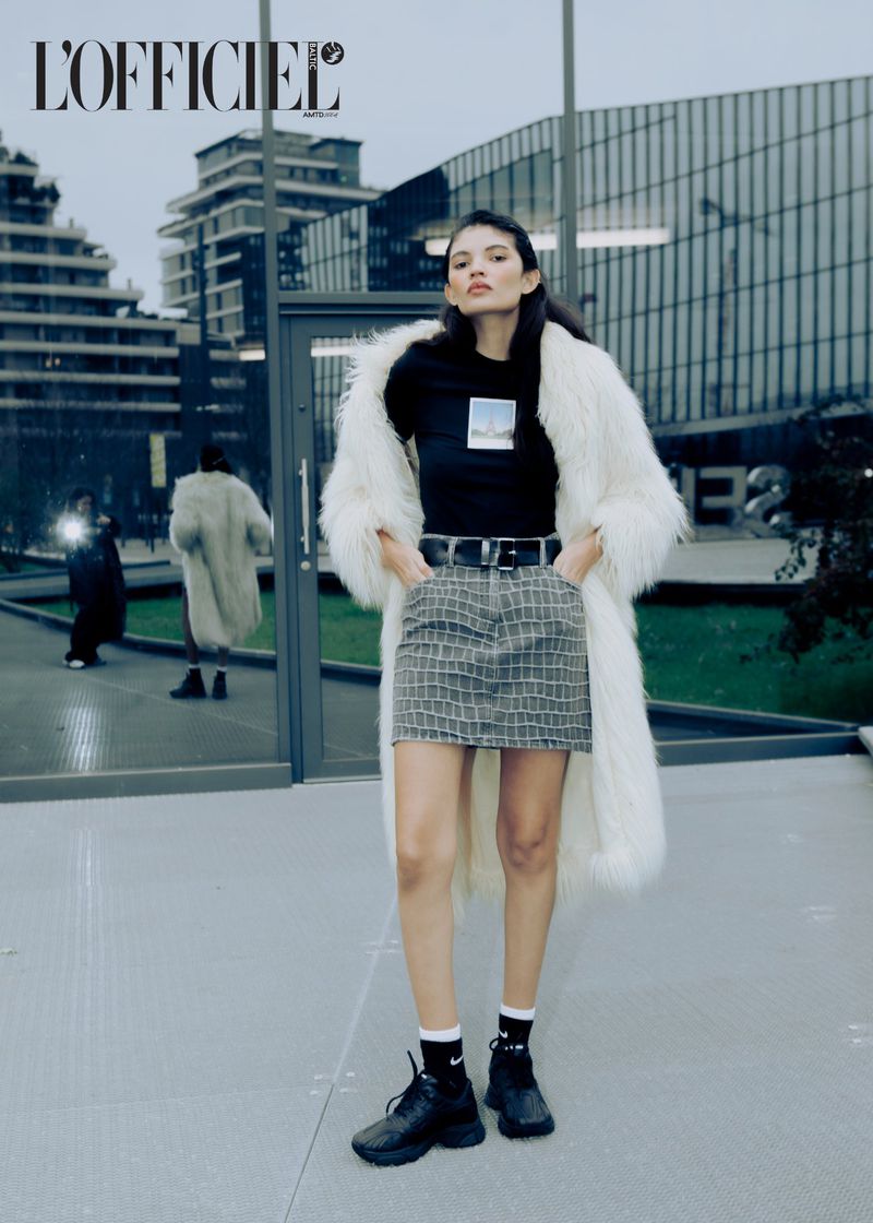 Fashion editorial image featuring a woman in a white faux fur coat, patterned skirt, and black top posing against a modern building.