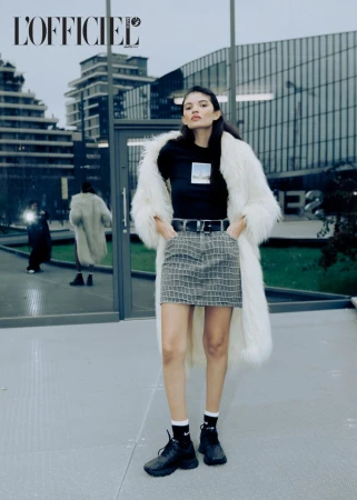 Fashion editorial image featuring a woman in a white faux fur coat, patterned skirt, and black top posing against a modern building.