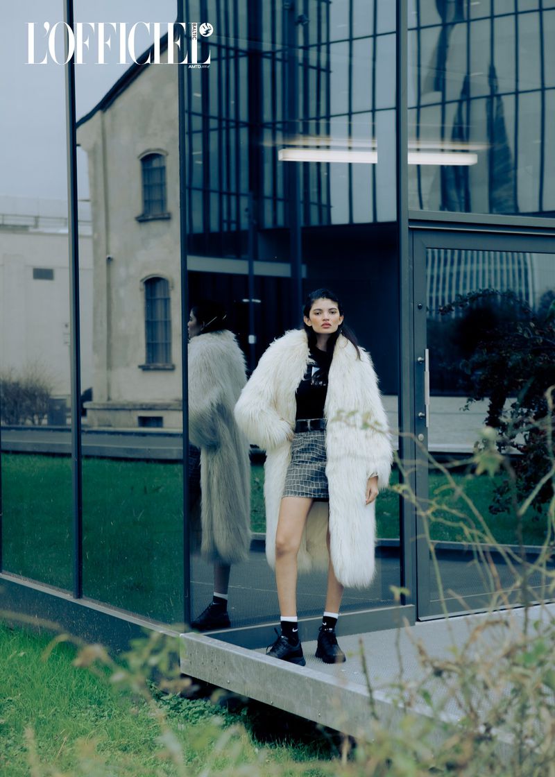 Fashion editorial image featuring a young woman in a white faux fur coat, mini skirt, and black boots, posing in front of a modern glass building.
