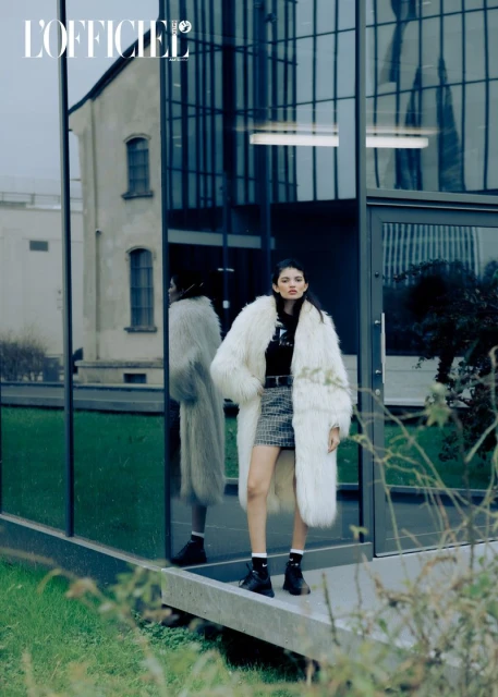 Fashion editorial image featuring a young woman in a white faux fur coat, mini skirt, and black boots, posing in front of a modern glass building.