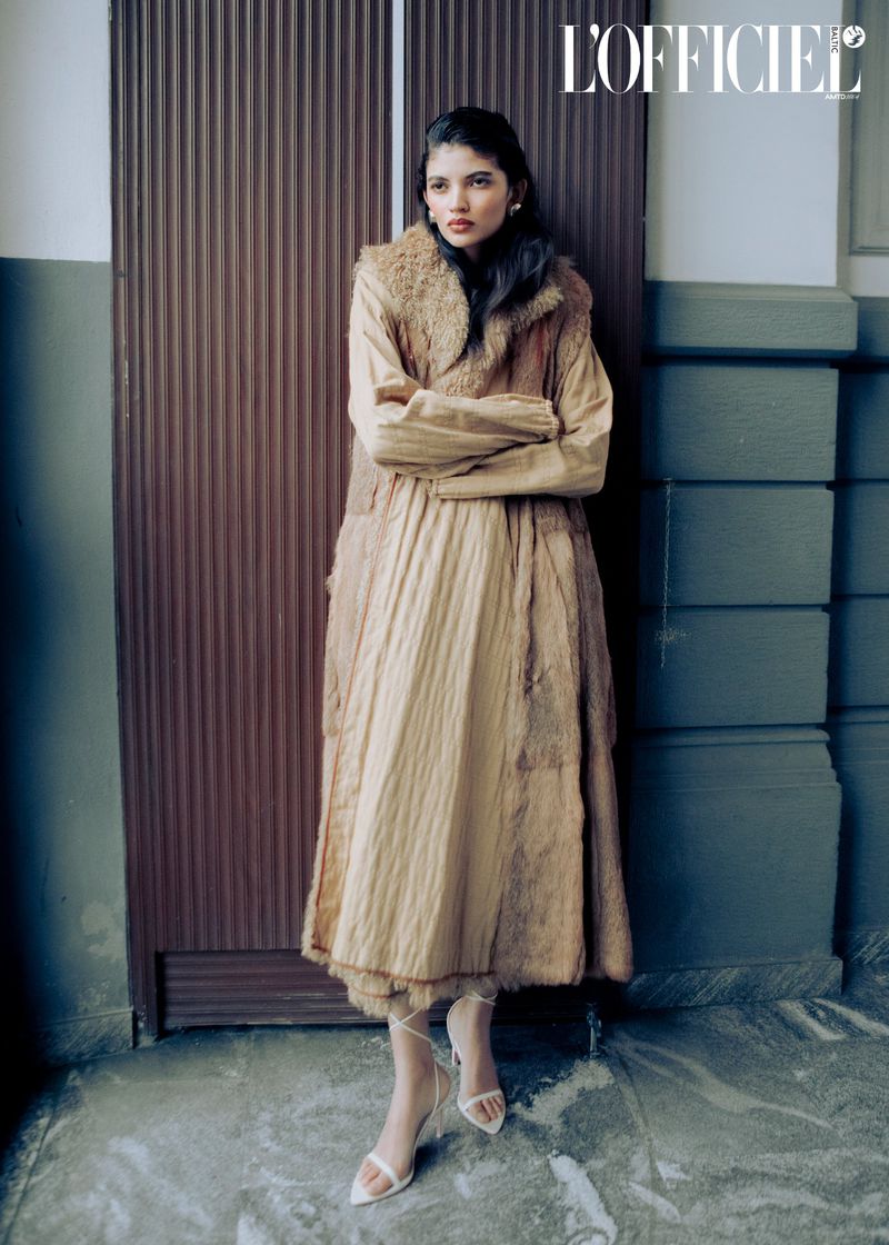 A woman poses in a long beige dress and fur vest in front of a grey wall and dark brown door for a fashion editorial.