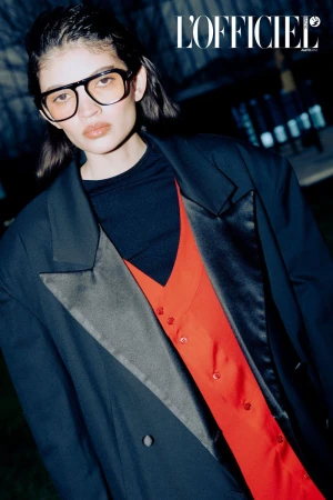 Woman wearing a black suit and glasses poses indoors for L'Officiel magazine, showcasing a fashion editorial style with dark lighting.