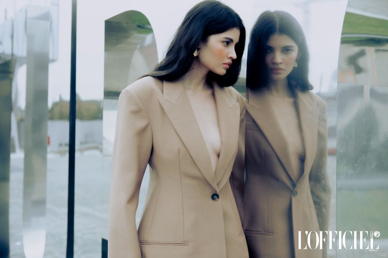 Brunette fashion model in a tan blazer poses near a mirrored surface in a stylish outdoor setting for a fashion magazine ad.