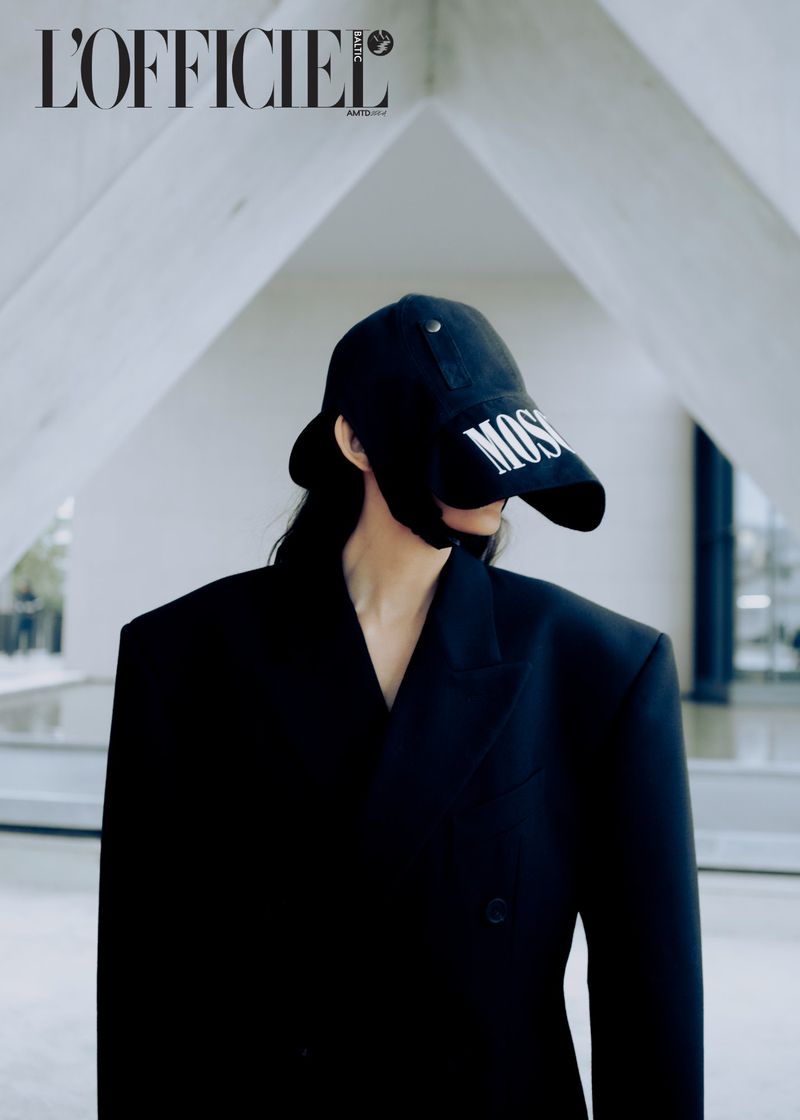 Woman in black blazer and cap stands in an outdoor architectural setting with a magazine logo in top left. Editorial fashion shot.