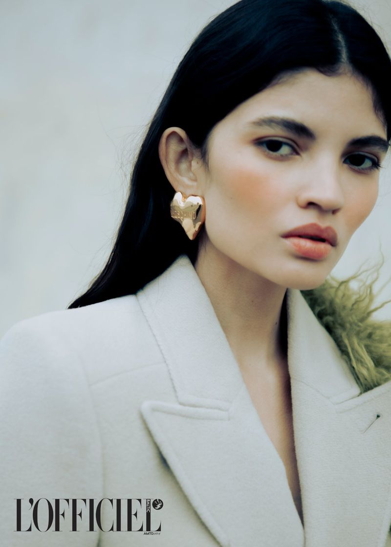 Close-up of a woman with dark hair wearing heart-shaped gold earrings and a cream-colored jacket, looking directly at the camera.