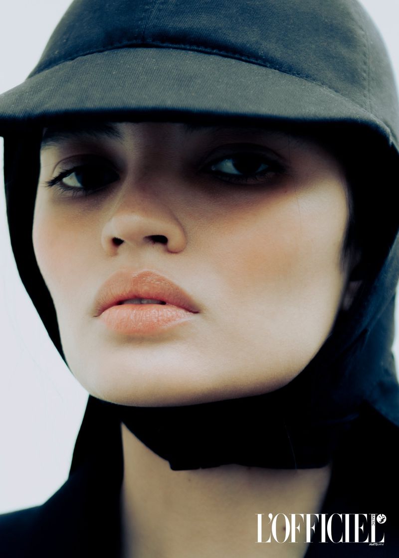 Close-up fashion portrait of a woman with dark hair wearing a black cap and jacket against a clean white background in a studio.