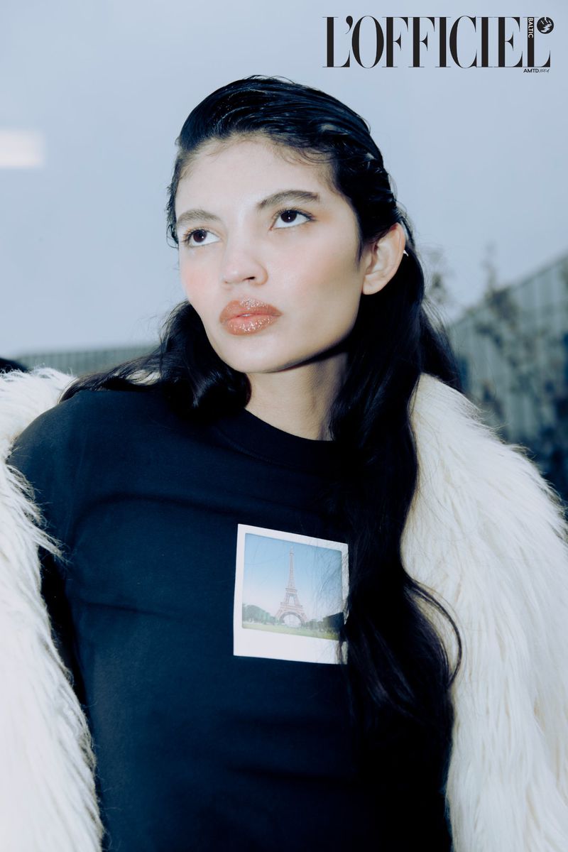 Fashion editorial portrait of a woman wearing a black shirt with Eiffel Tower graphic and a white fur coat, looking up outdoors.