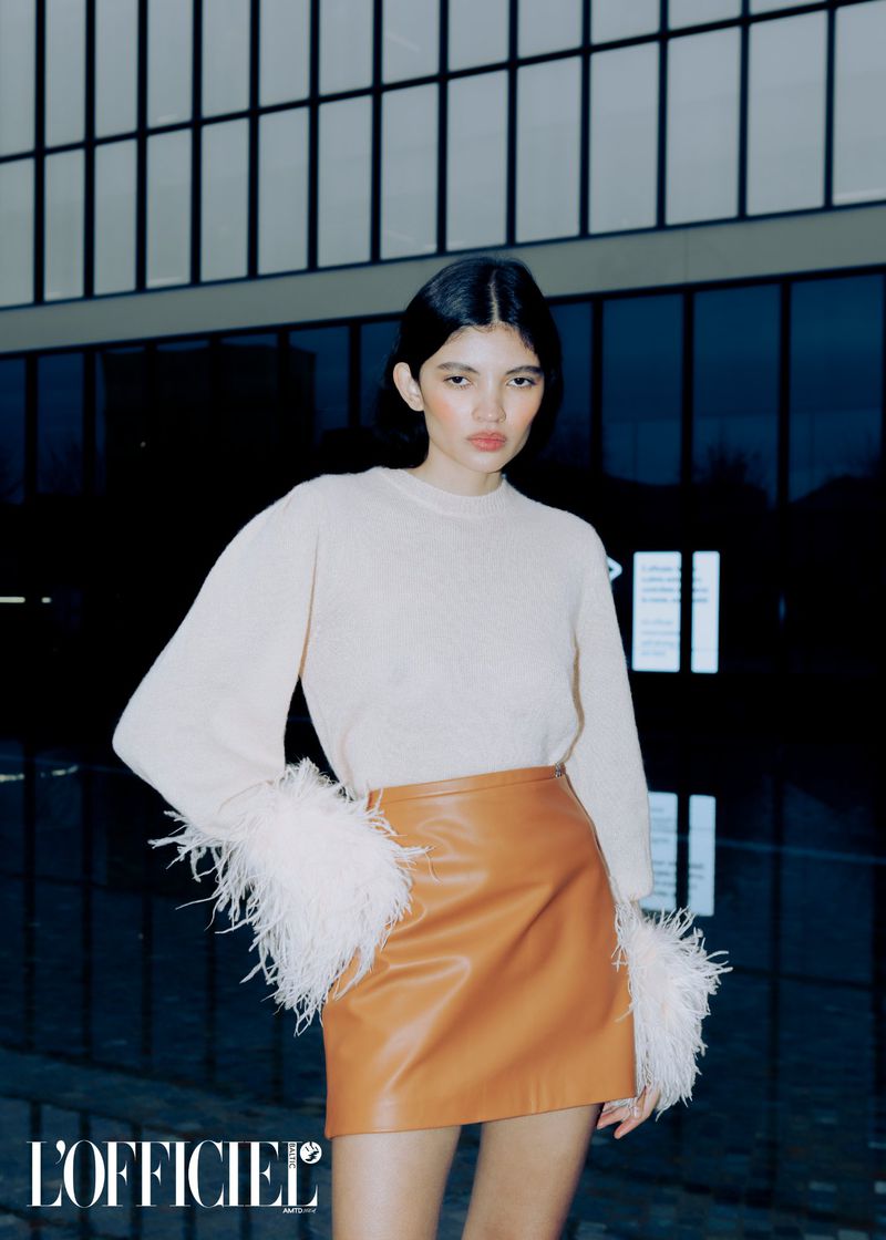 Fashion shot of a model wearing a beige top with feather cuffs and a tan leather miniskirt, posing against an architectural backdrop.