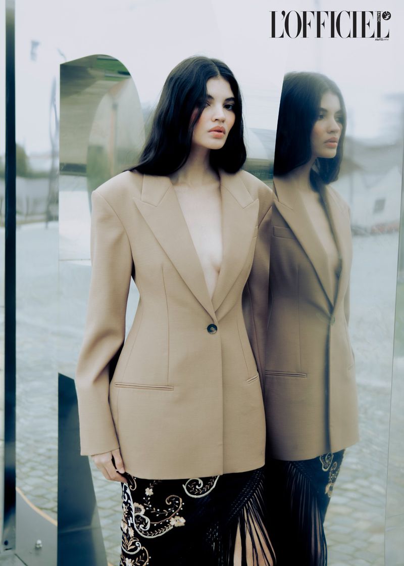 Fashion editorial shot of a brunette adult woman in a beige blazer and patterned skirt standing next to a reflective surface outdoors.