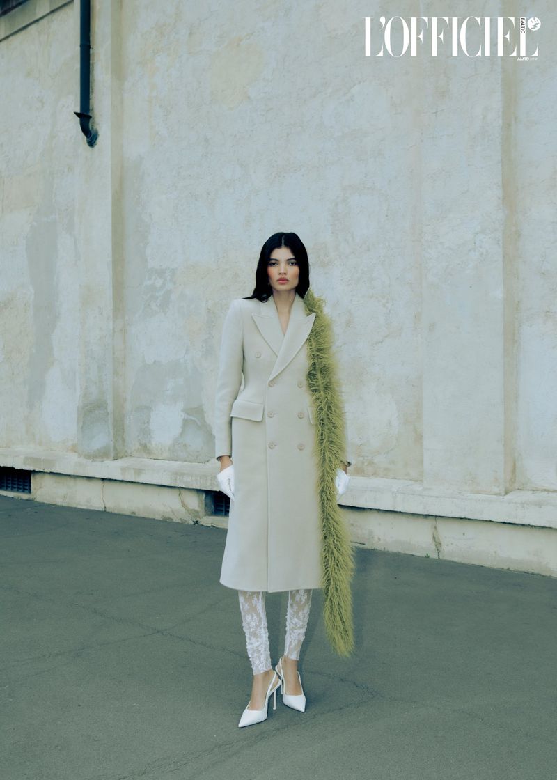 A stylish woman in a beige coat and patterned leggings stands against a textured wall in a fashion editorial shot with natural light.