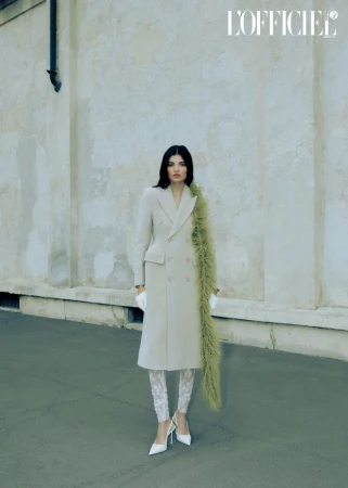 A stylish woman in a beige coat and patterned leggings stands against a textured wall in a fashion editorial shot with natural light.