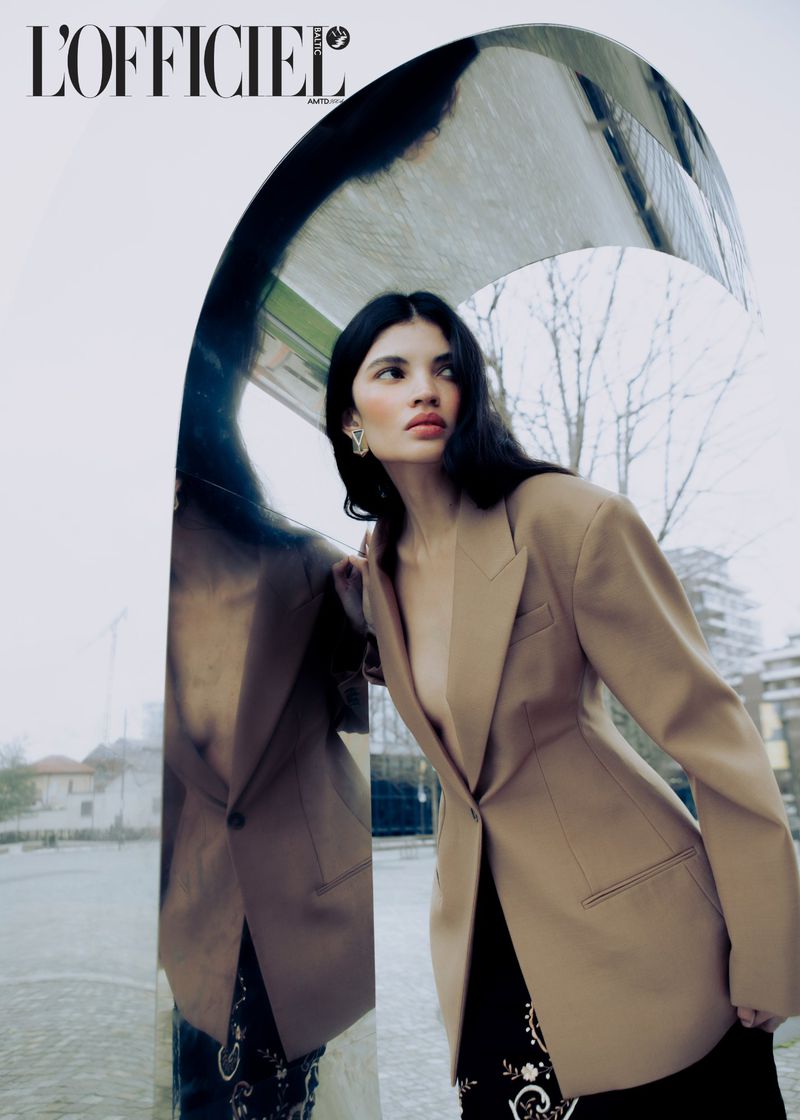 Fashion editorial image featuring a female model in a blazer posing against a reflective arched wall with trees and buildings visible.