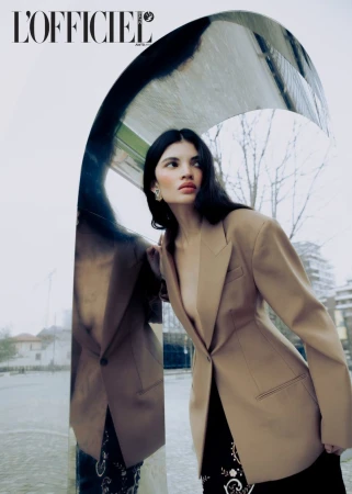 Fashion editorial image featuring a female model in a blazer posing against a reflective arched wall with trees and buildings visible.