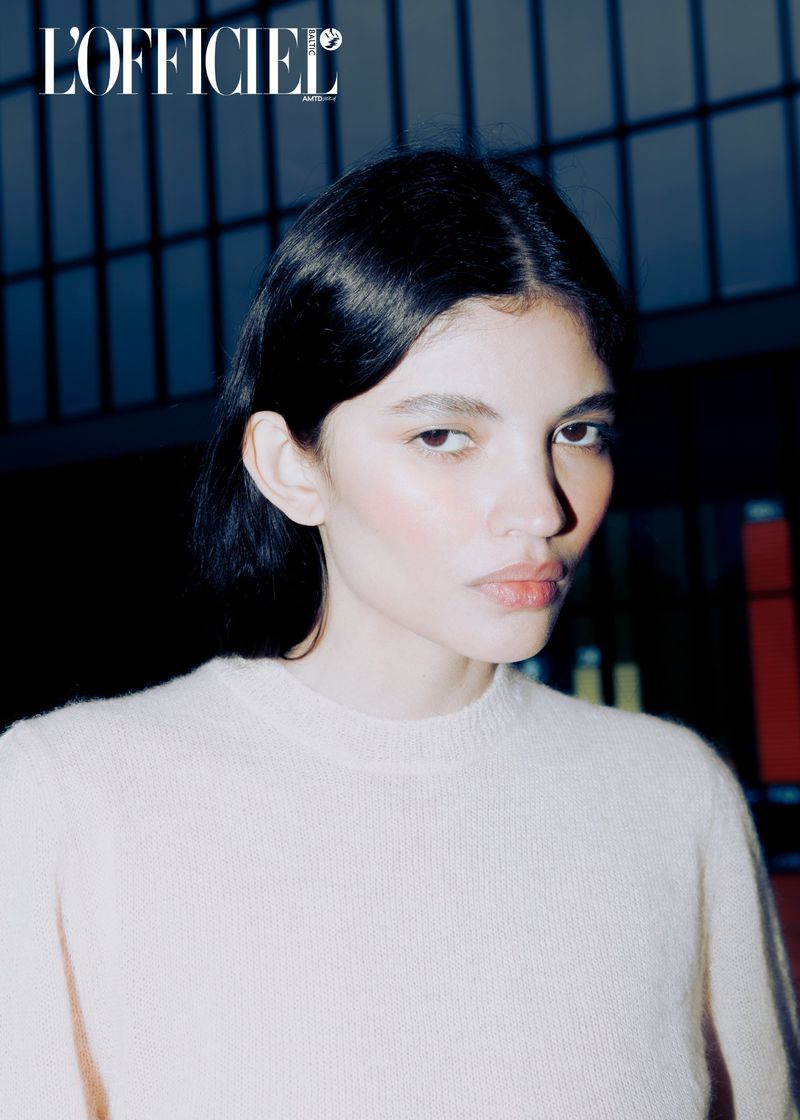 Close-up portrait of a young woman with dark hair wearing a pale sweater, set against a dark background. Magazine logo visible.