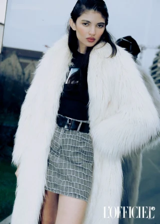 Fashion editorial shot of a woman wearing a faux fur coat, black top, and grey mini skirt, posing in front of a reflective glass building.