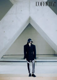 Model wearing oversized black blazer, baseball cap, and patterned leggings standing beneath concrete architecture