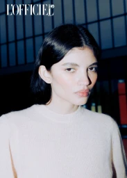 Close-up portrait of a woman with dark hair wearing a light-coloured knitted sweater against an indoor window background.