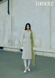 Model in cream double breasted coat with green feather boa standing against a textured rustic wall for L Officiel Baltics editorial