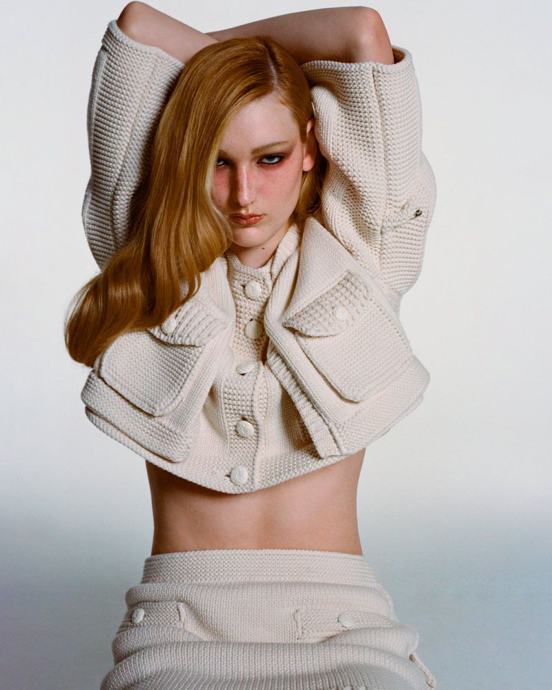 Blonde-haired fashion model wearing a knitted jacket and skirt, posing with arms raised in a studio setting against a neutral background.