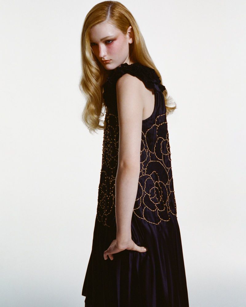 A fair-skinned young woman with long blonde hair poses in a sleeveless black dress with gold floral details against a white backdrop.
