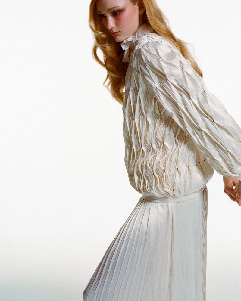 A fair-skinned model with long blonde hair poses against a white background, wearing a pleated white top and flowing white skirt.