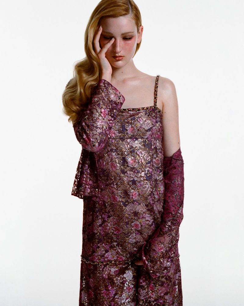 A fair-skinned fashion model in a floral-patterned dress and lace gloves poses on a white background, with her hand near her face.