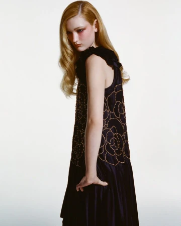 Model with long blonde hair wearing a black sleeveless dress with gold embroidery against a bright white background