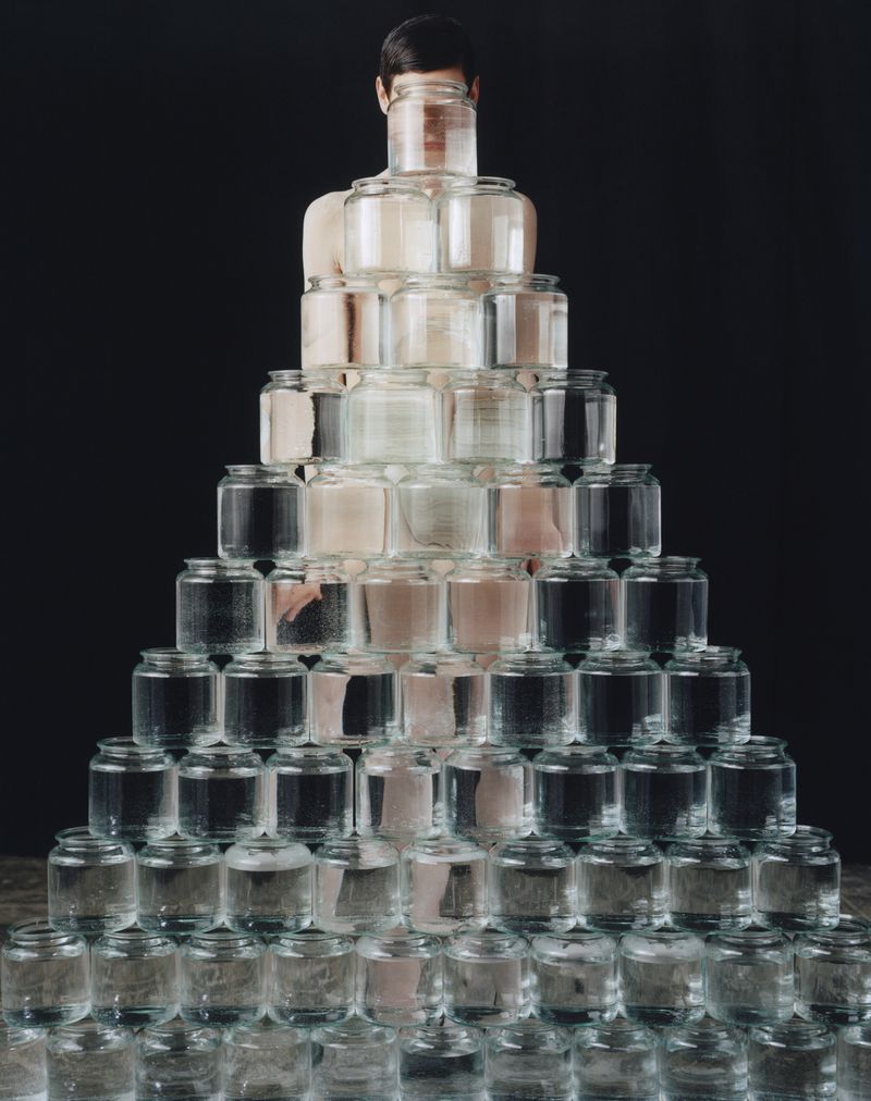 Creative studio shot of a person layered in glass jars of differing sizes, stacked in a triangular shape on a wooden surface.