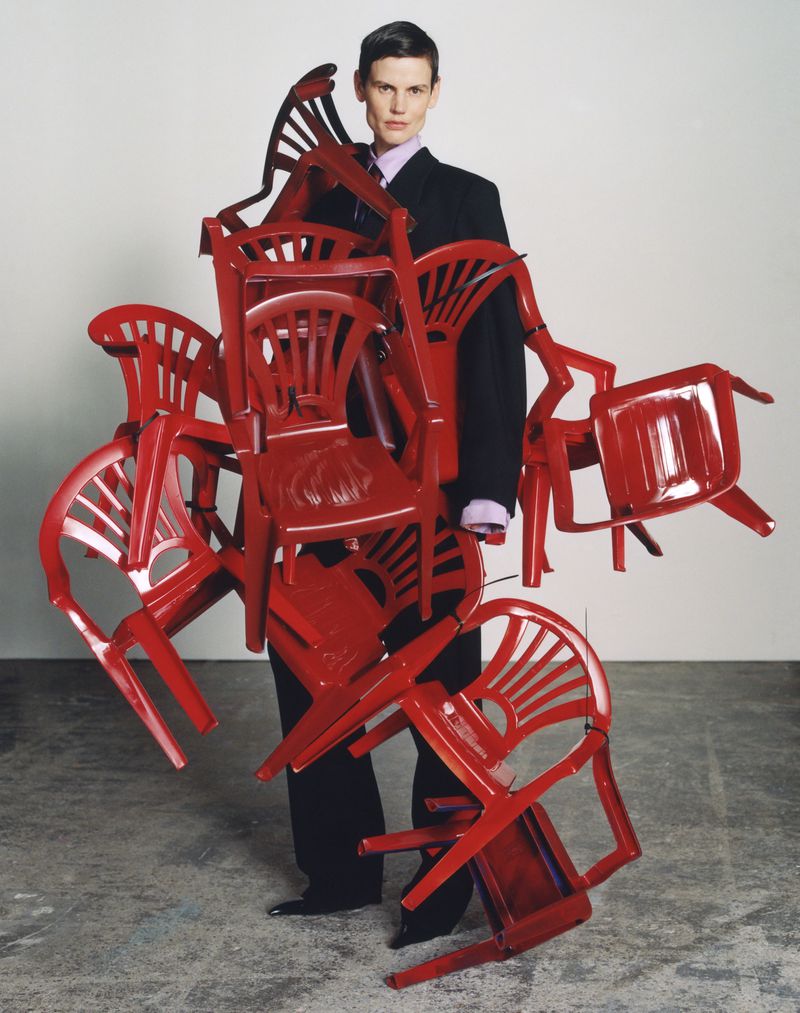 Adult standing while covered in red plastic chairs in a studio, wearing a black suit and looking at the camera.