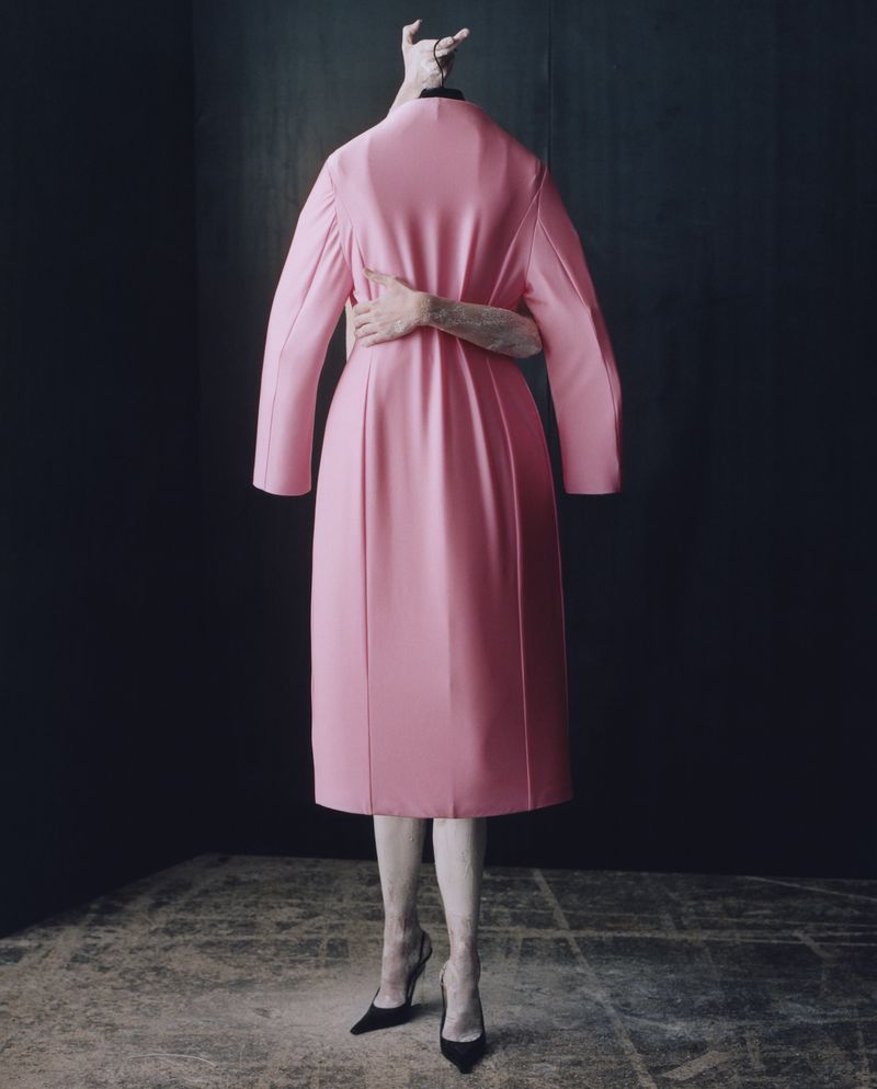Surreal fashion image featuring a pink coat worn by an invisible person, posed with visible legs in heels and a hand on the coat.