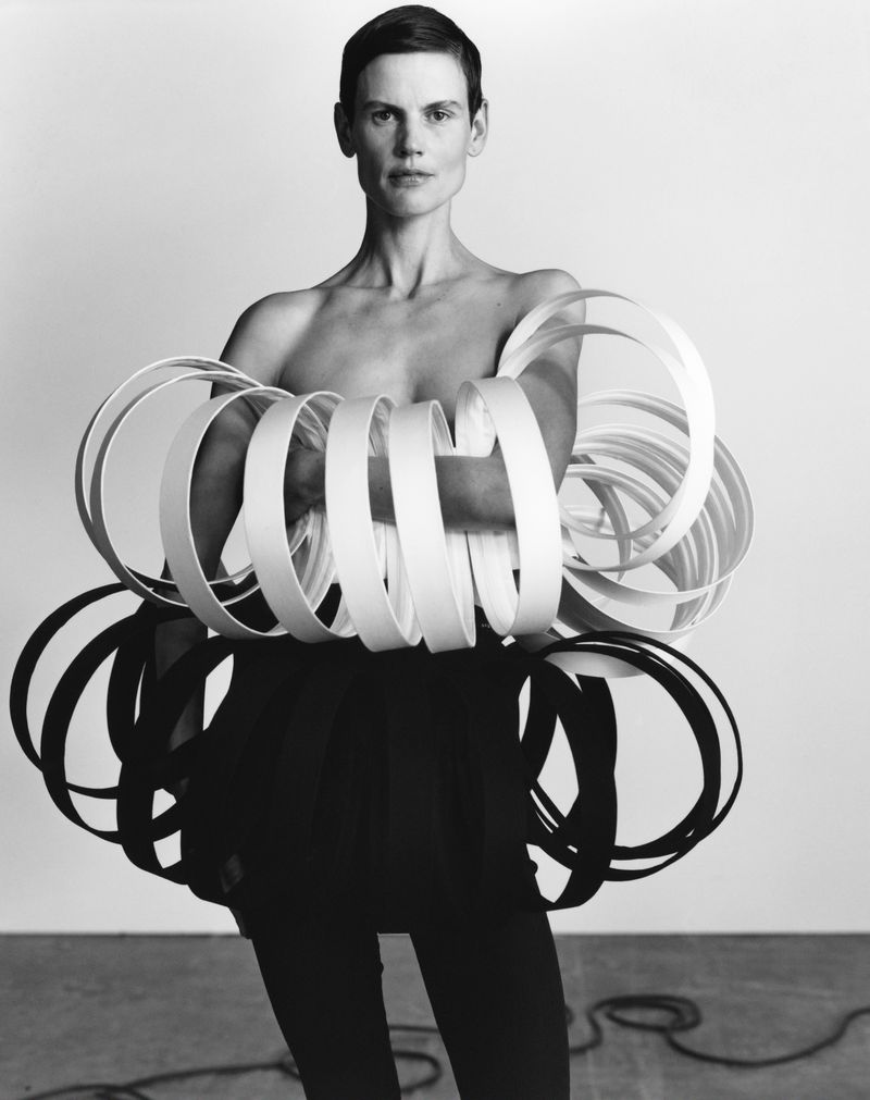 Black and white studio portrait of a bare-chested person surrounded by white and black concentric rings, wearing dark pants.