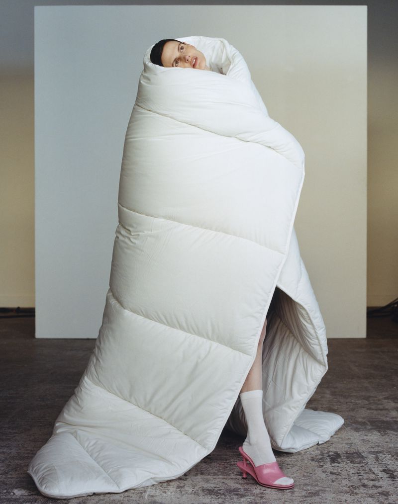 A woman posing indoors wrapped in a puffy white comforter, wearing white socks and pink high-heeled shoes, in a studio setting.