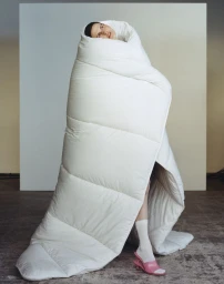 Model wrapped entirely in a puffy white duvet with only face and legs visible against a neutral background