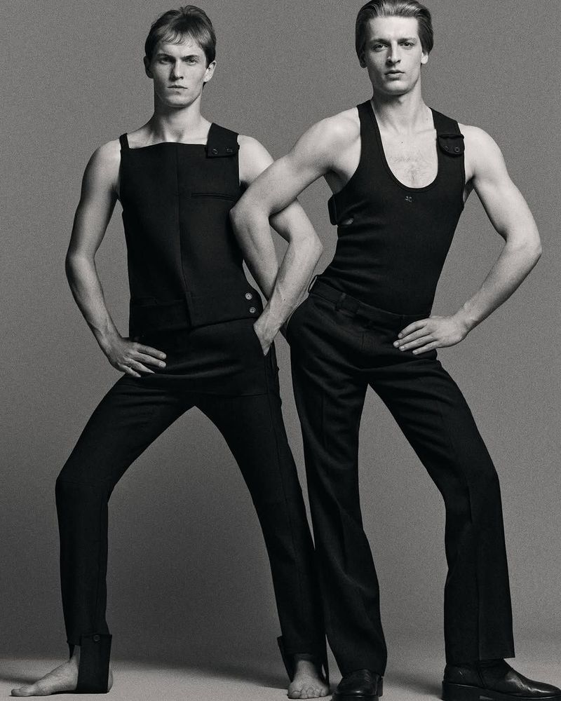 Two male models in black tank tops and pants pose in a studio with hands on hips, one barefoot, the other in dress shoes, against a grey backdrop.