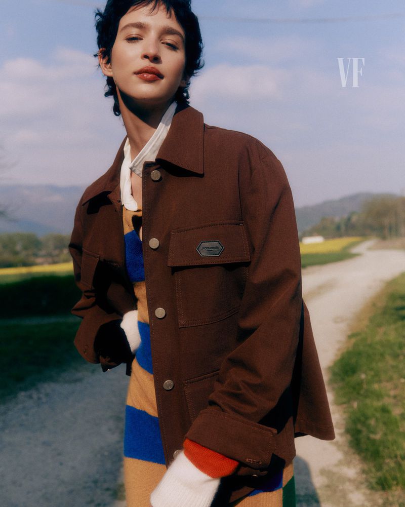 Woman in a brown jacket stands on a rural road with a colorful striped sweater on a sunny day, fashion editorial with VF logo.