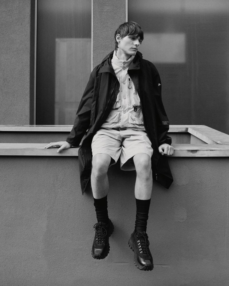 Monochrome shot of a slender male model seated outdoors on a ledge, wearing a dark coat, khaki shorts, dark socks, and lug-soled shoes.