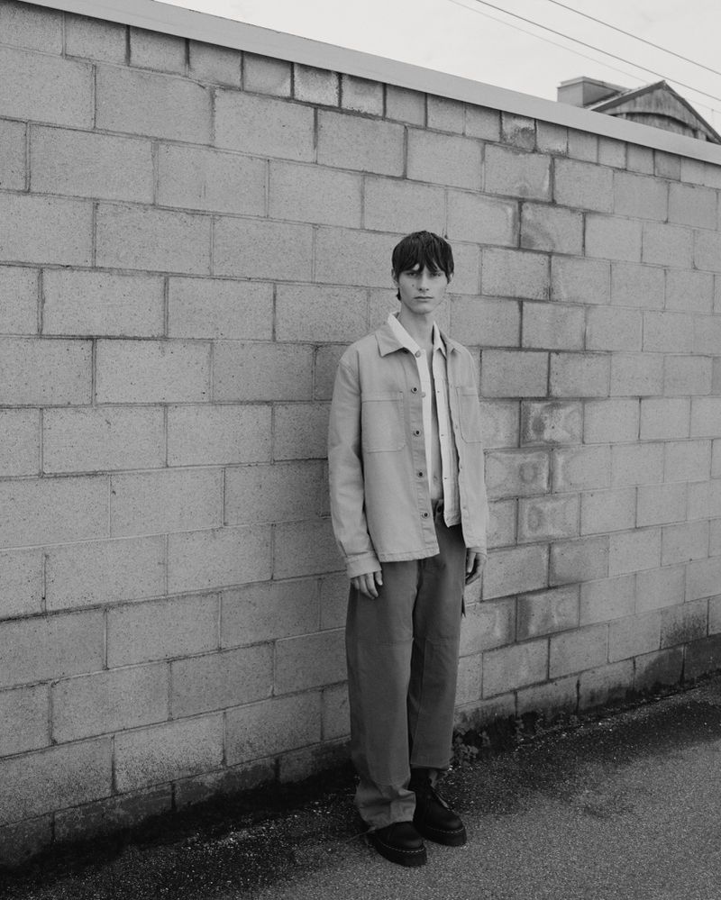 Adult male in casual jacket and trousers stands against a cinder block wall outdoors, shot in black and white with overcast lighting.