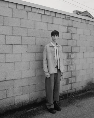 Adult male in casual jacket and trousers stands against a cinder block wall outdoors, shot in black and white with overcast lighting.