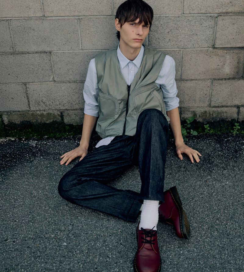 Young adult poses outdoors against a brick wall wearing a vest, button-down shirt, denim jeans, burgundy shoes, and white socks.
