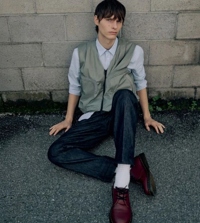 Young adult poses outdoors against a brick wall wearing a vest, button-down shirt, denim jeans, burgundy shoes, and white socks.